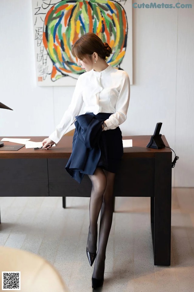 A woman sitting at a desk in a white shirt and black skirt.